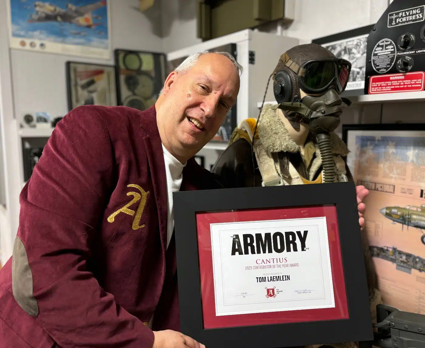 Tom Laemlein in the crimson Cantius Award jacket, holding his framed Cantius Award certificate surrounded by aviation memorabilia.